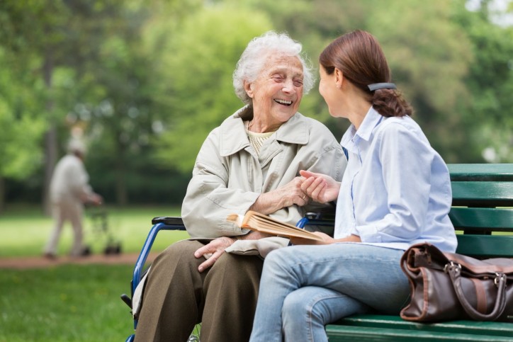 Eine Seniorin sitzt mit einer etwa 40 jährigen Frau auf einer Bank. Beide reichen sich die Hände und lächeln sich an.