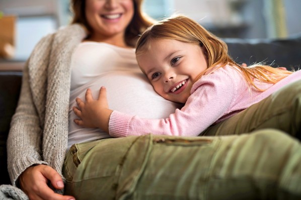 Ein etwa 5-jähriges Kind liegt mit dem Kopf auf dem Babybauch einer sitzenden Frau.