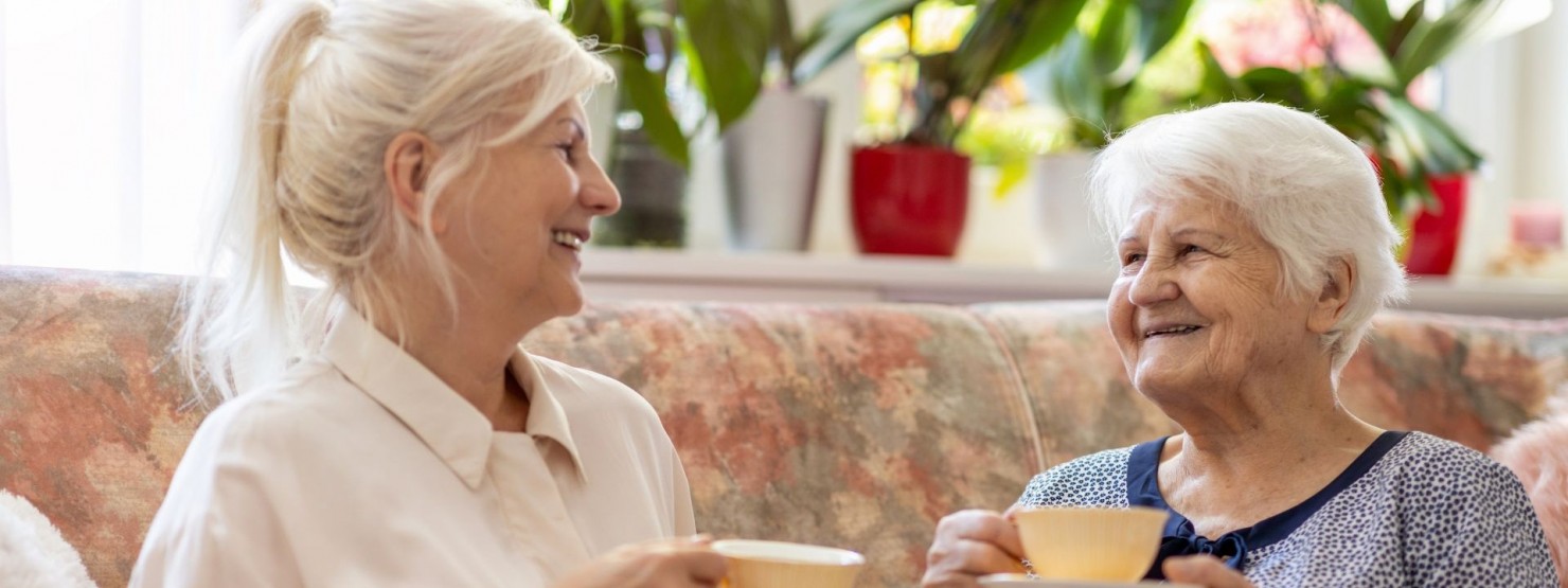 Zwei Seniorinnen sitzen auf einer Couch im Wohnzimmer und trinken gemeinsam Kaffee. Beide sind zueinander gedreht und lächeln sich an.