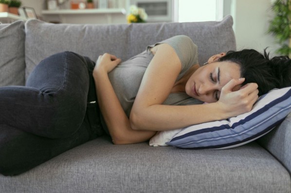 Junge Frau liegt auf ihrer Couch. Sie krümmt sich vor Schmerzen im Bauch. 