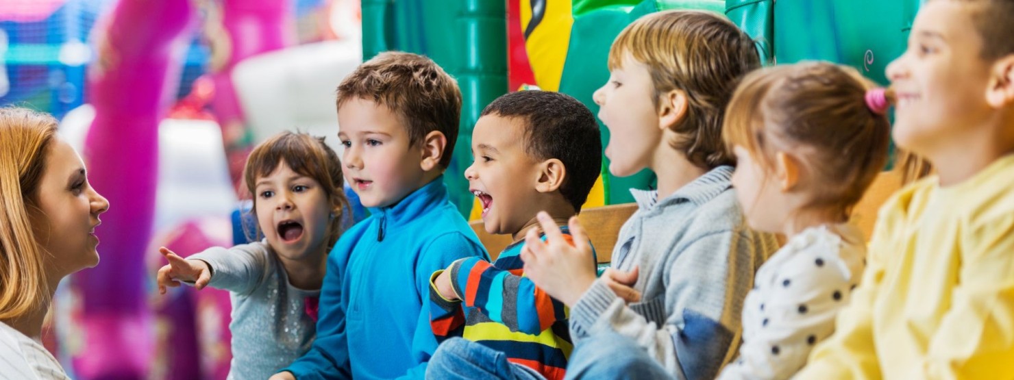Eine Kindergruppe sitzt zusammen auf einer Bank und lacht zusammen. Eine Erzieherin hockt vor ihnen und spricht.