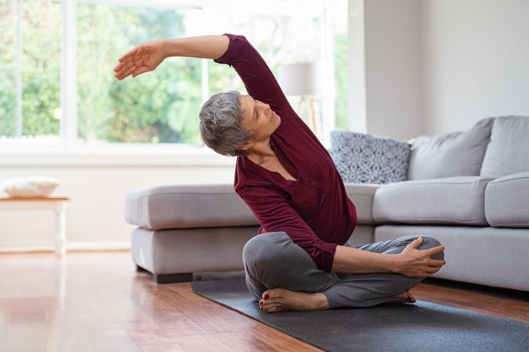 Eine etwa 60 jährige Frau sitzt im Schneidersitz auf einer Yogamatte im Wohnzimmer. Sie dehnt ihren linken Arm über ihren Kopf hinweg nach rechts.