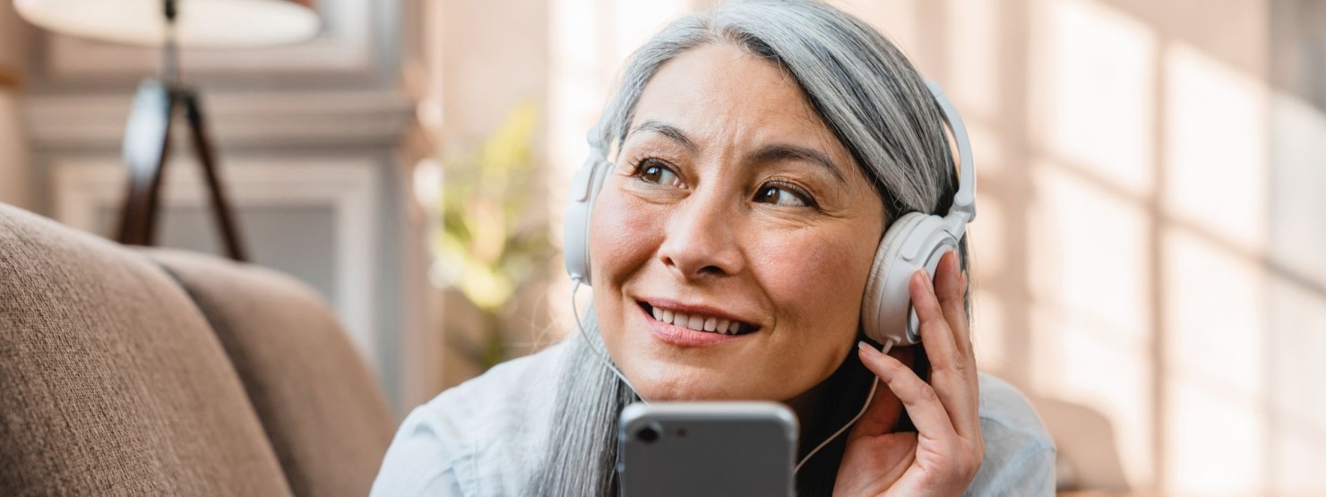 Eine Seniorin liegt auf einem Sofa und hört über Kopfhörer den AOK-Podcast "ausgesprochen menschlich - Selbsthilfe auf Sendung".