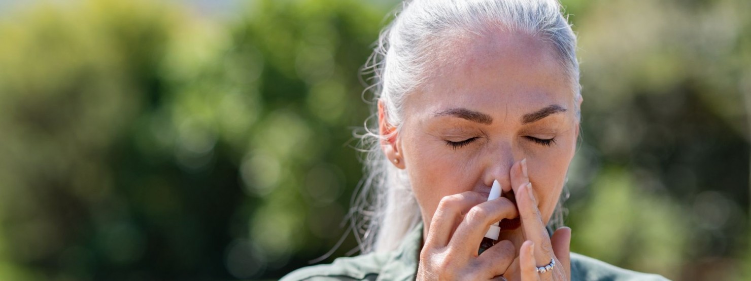Eine etwa 50 jährige Frau steht in der Natur und hat die Augen geschlossen. Sie hat beide Hände im Gesicht und führt sich ein Nasenspray in die Nase ein.