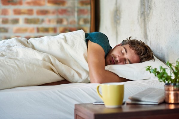 Ein Mann Mitte 30 liegt in seinem Bett und schläft. Auf seinem Nachttisch steht eine Tasse Tee. 