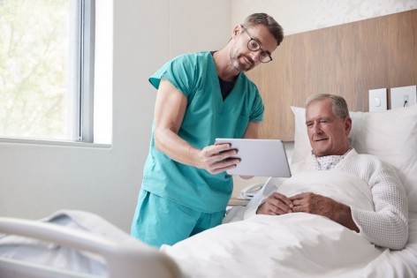 Ein Krankenpfleger steht am Bett eines Patienten und zeigt etwas auf einem Tablet.