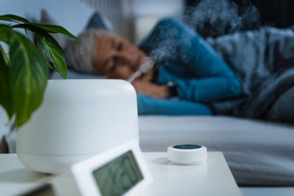 Eine etwa 50 jährige schläft in einem Bett. Im Vordergrund steht auf einem Nachttisch ein aktiver Luftbefeuchter und ein Wecker.