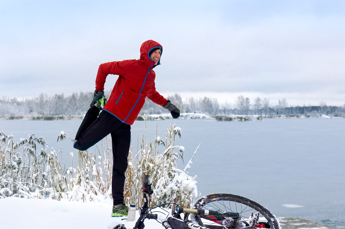 Ein etwa 30-jähriger Mann mit dicker Jacke macht im Winter an einem See Gymnastik um sich aufzuwärmen. Vor ihm liegt sein Fahrrad im Schnee.