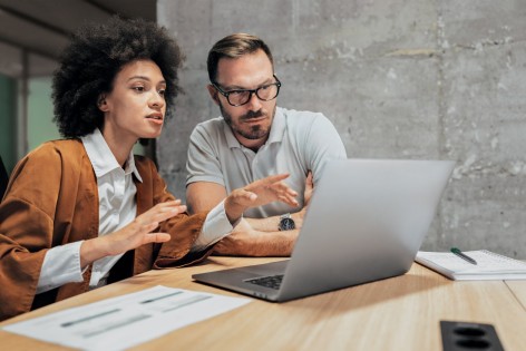 Eine Frau mit schwarzen Locken erklärt einem Mann, beide etwa 35, etwas am Laptop. Er trägt eine Brille und ein weißes T-Shirt, sie eine weiße Bluse und eine braune Jacke.