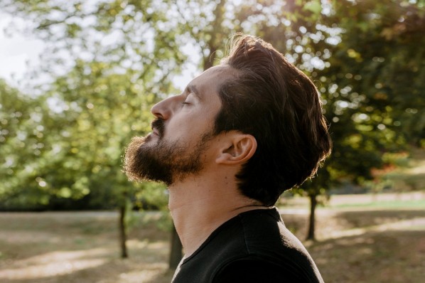 Ein Mann Mitte 30 mit einem Vollbart schließt die Augen und lehnt seinen Kopf zurück, während die Sonne ihn von der Seite bescheint.