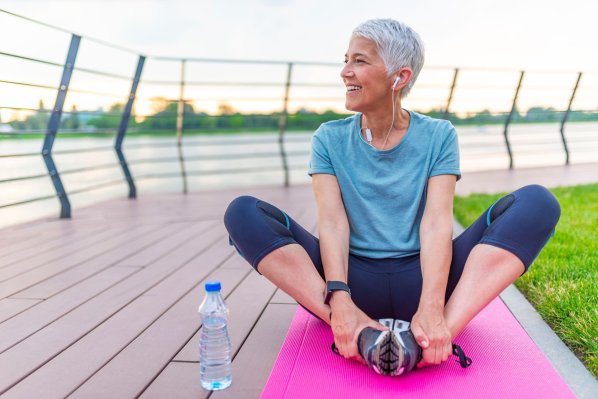 Eine etwa 60 jährige Frau sitzt im Schneidersitz auf einer Sportmatte am Rande eines Sees. Neben ihr steht eine kleine Wasserflasche. Sie schaut lächelnd aufs Wasser und dehnt dabei ihre Beine.