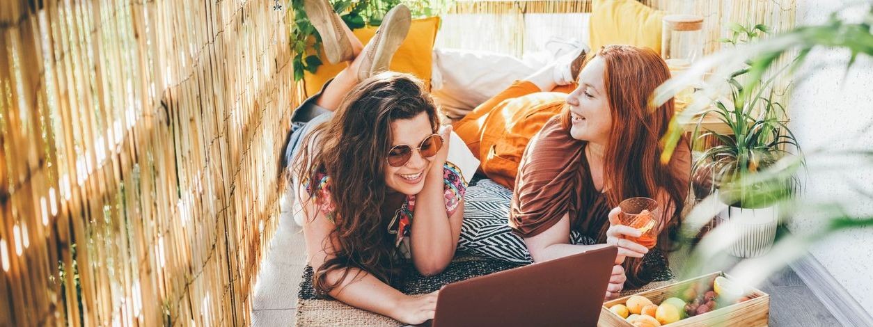 Zwei junge Frauen liegen auf einem Balkon in der Sonne, lachen und schauen auf einen Laptop.