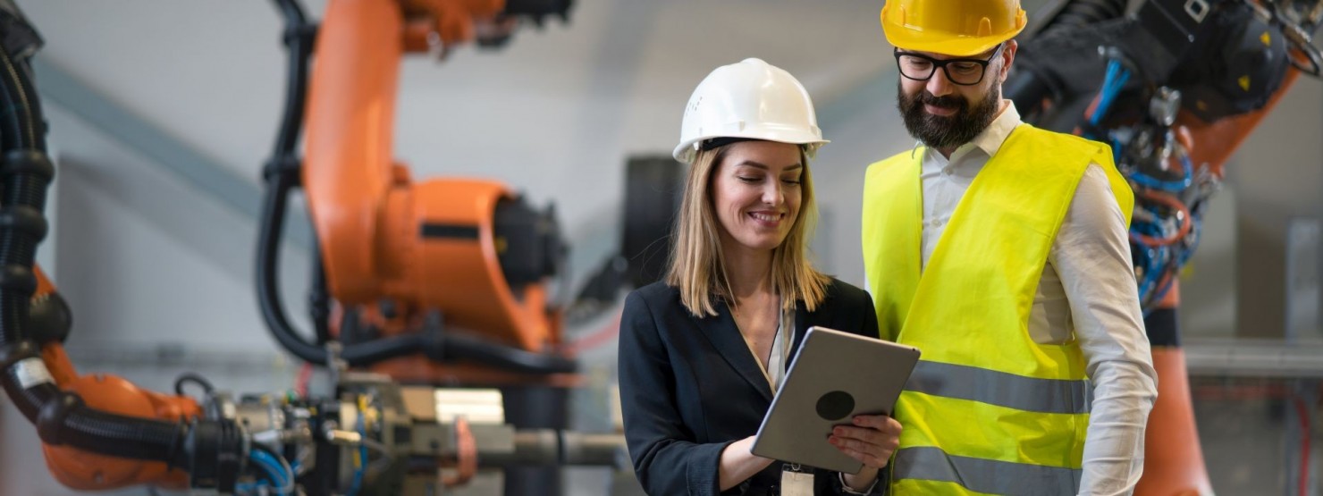 Eine Frau und ein Mann stehen neben einer Arbeitsmaschine. Bei tragen eine Helm. Die Frau zeigt etwas auf dem Tablet.