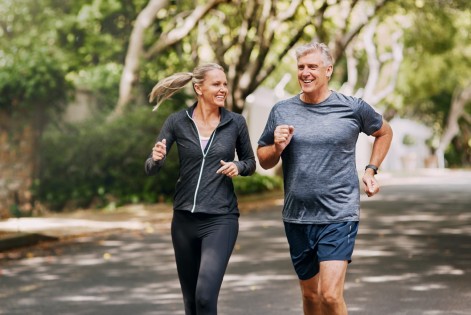 Ein Mann und eine Frau, beide circa Mitte 50, joggen gemeinsam eine von Bäumen umsäumte Straße entlang. Beide strahlen Entspannung und Energie aus. 