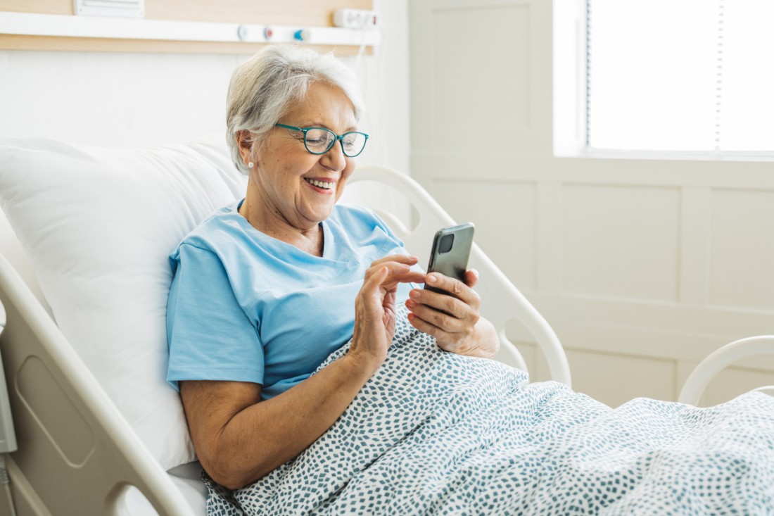 Seniorin liegt im Krankenhausbett. Sie lächelt und schaut auf ihr Smartphone