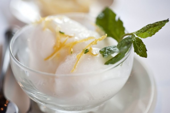 Auf einem weißen Teller steht eine durchsichtige Schale gefüllt mit Zitronensorbet. Es ist dekoriert mit einem Minzblatt und Zitronenschalen.