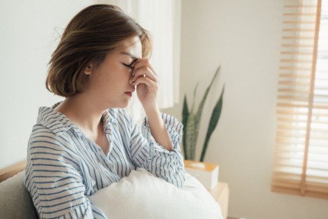 Junge Frau sitzt übermüdet in ihrem Bett. Sie hält ihren Kopf vor Müdigkeit fest. 