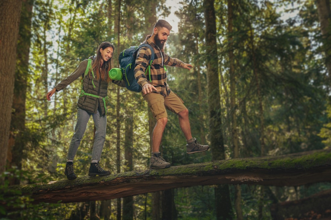 Ein Pärchen balanciert im Wald über einen umgestürtzen Baum. Sie sind beide etwa 30 Jahre alt und tragen Wandersachen und einen Rucksack.