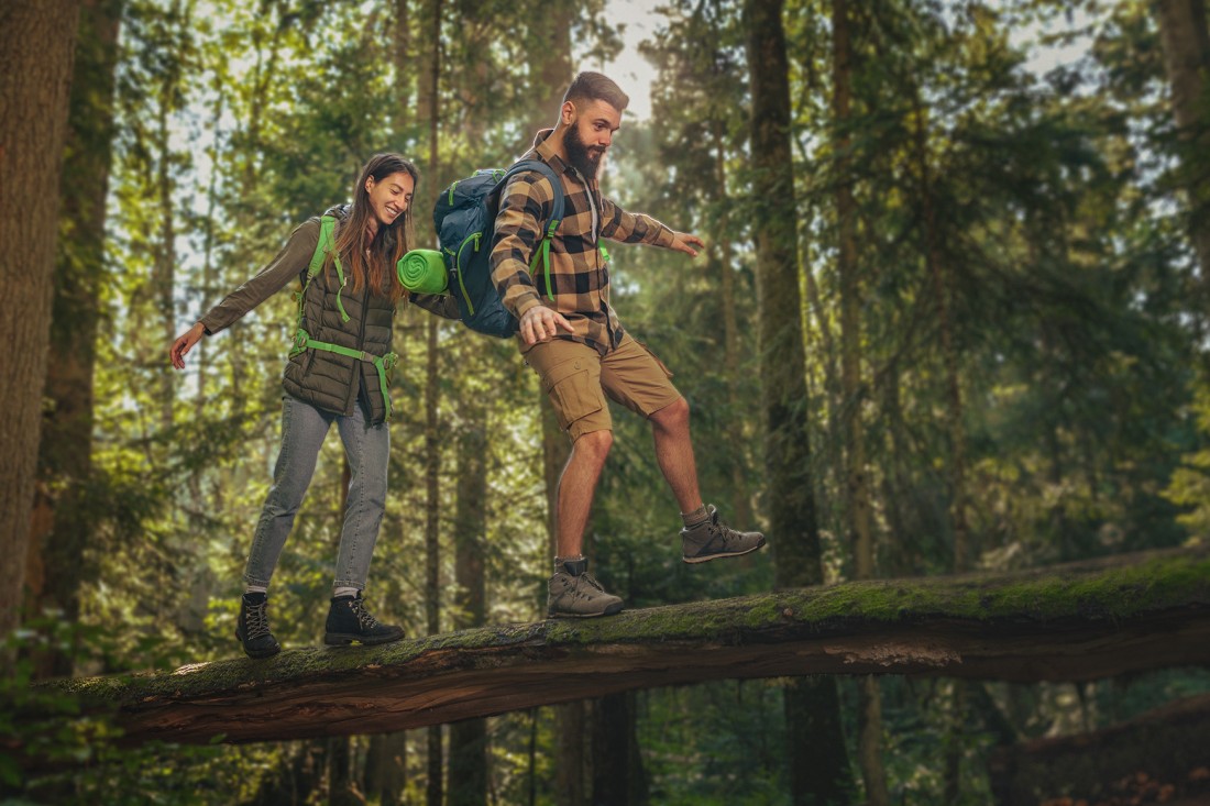 Ein Pärchen balanciert im Wald über einen umgestürtzen Baum. Sie sind beide etwa 30 Jahre alt und tragen Wandersachen und einen Rucksack.