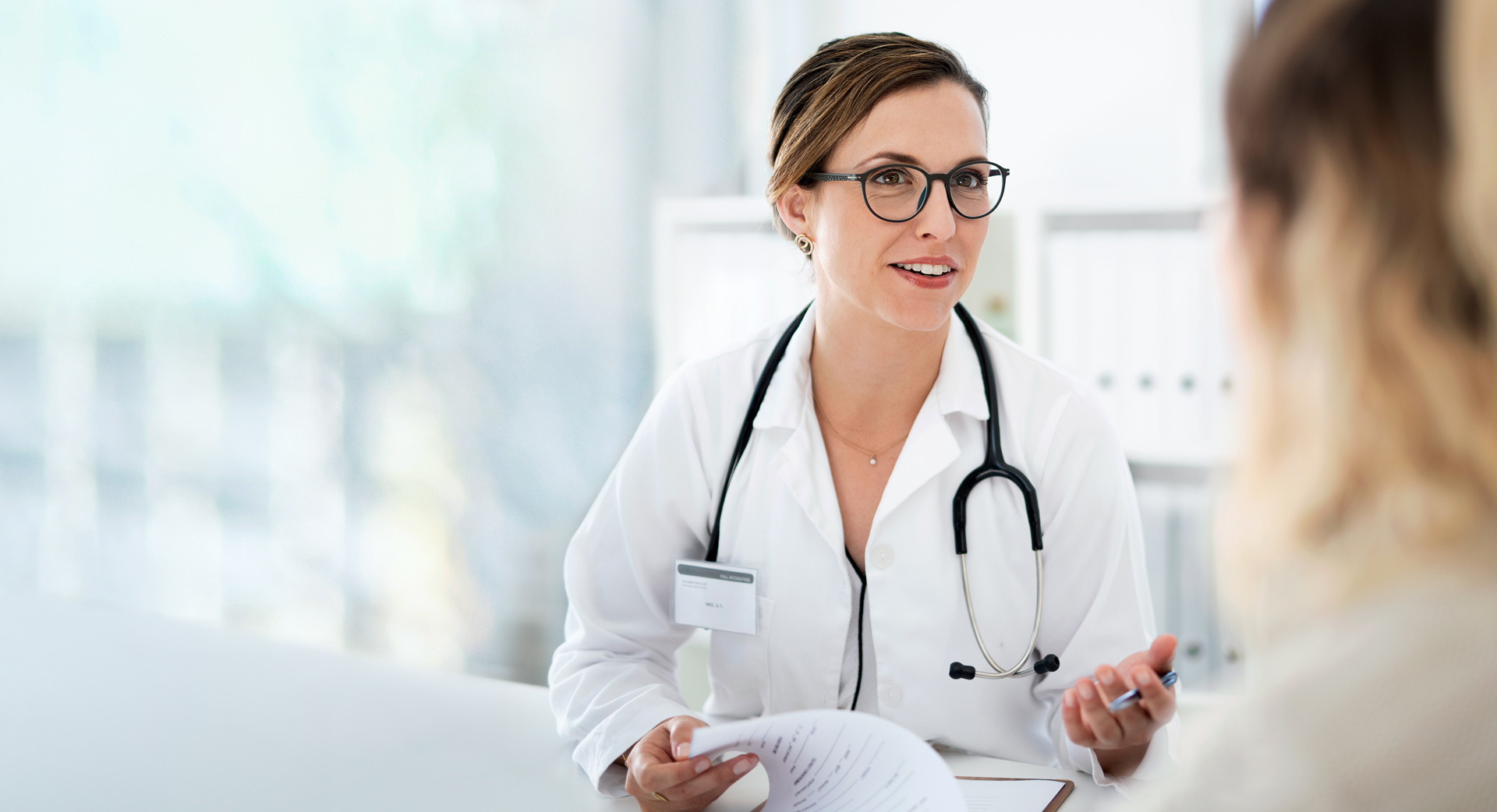 Eine Ärztin sitzt an einem Tisch und berät einen Patienten. Auf dem Tisch stehen ein Laptop und eine Packung Tabletten.