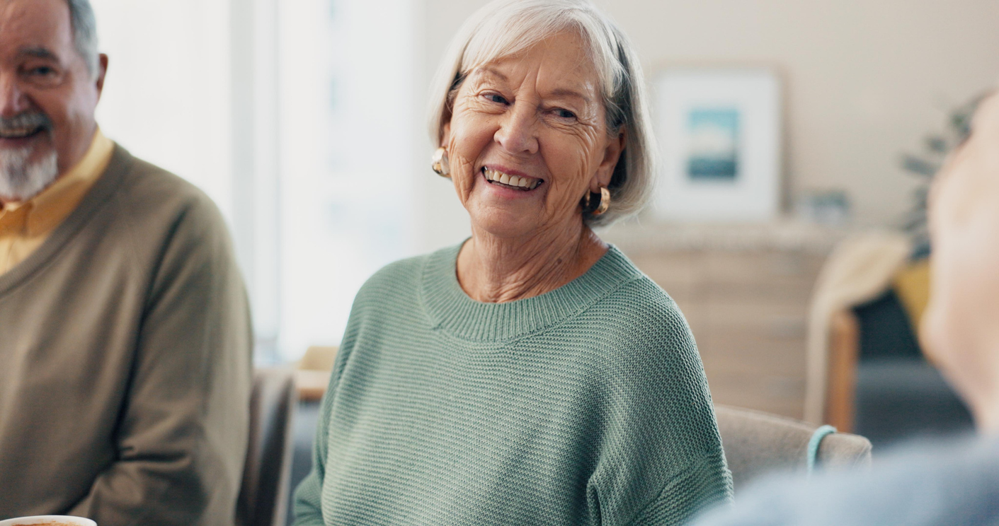 Zwei Seniorinnen sitzen auf einer Couch im Wohnzimmer und trinken gemeinsam Kaffee. Beide sind zueinander gedreht und lächeln sich an.