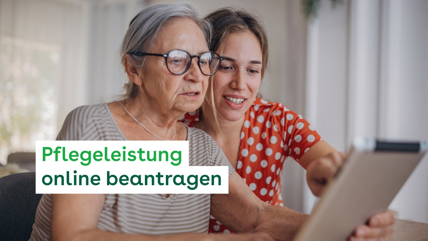 Zwei Frauen, eine etwa 30 Jahre und eine etwa 80 Jahre, sitzen zusammen an einem Tisch. Die ältere Frau hält ein Tablet und blickt durch ihre Brille interessiert darauf. Die jüngere lehnt von hinten leicht über ihre Schulter und zeigt auf etwas auf dem Tablet.