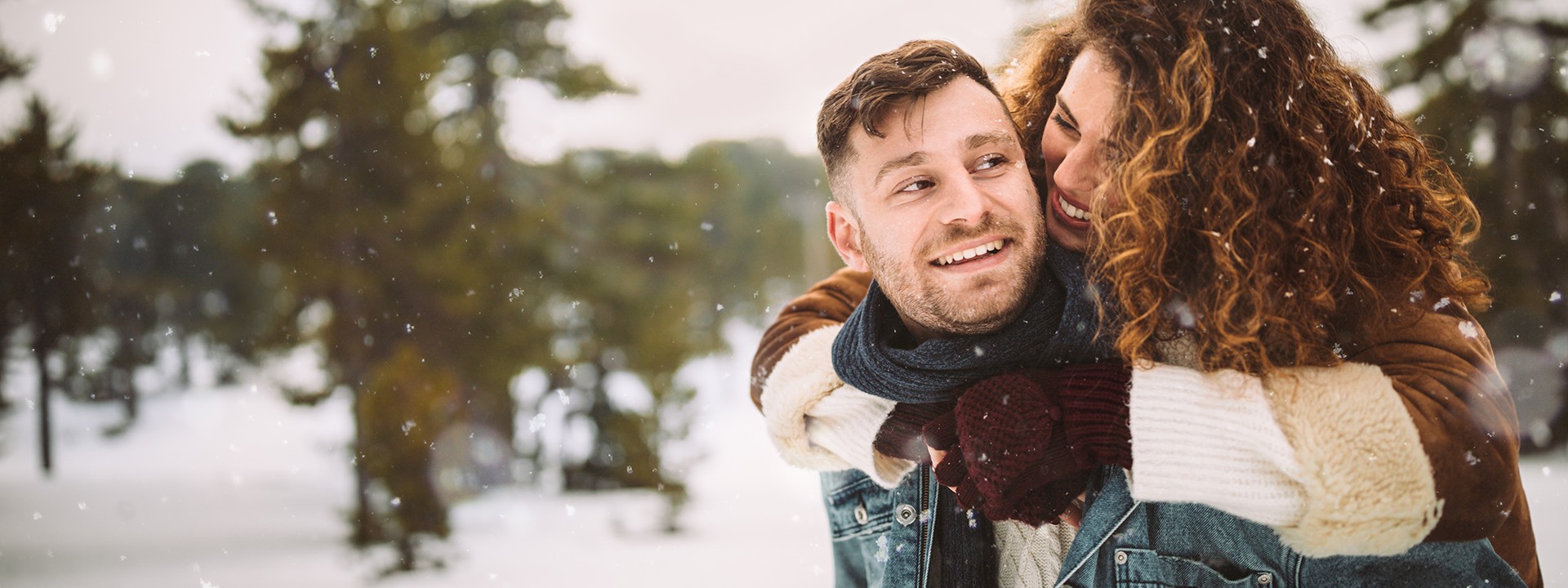 Ein etwa 35-jähriger Mann trägt seine etwa 30-jährige Frau auf dem Rücken durch den Winterwald. Es schneit.
