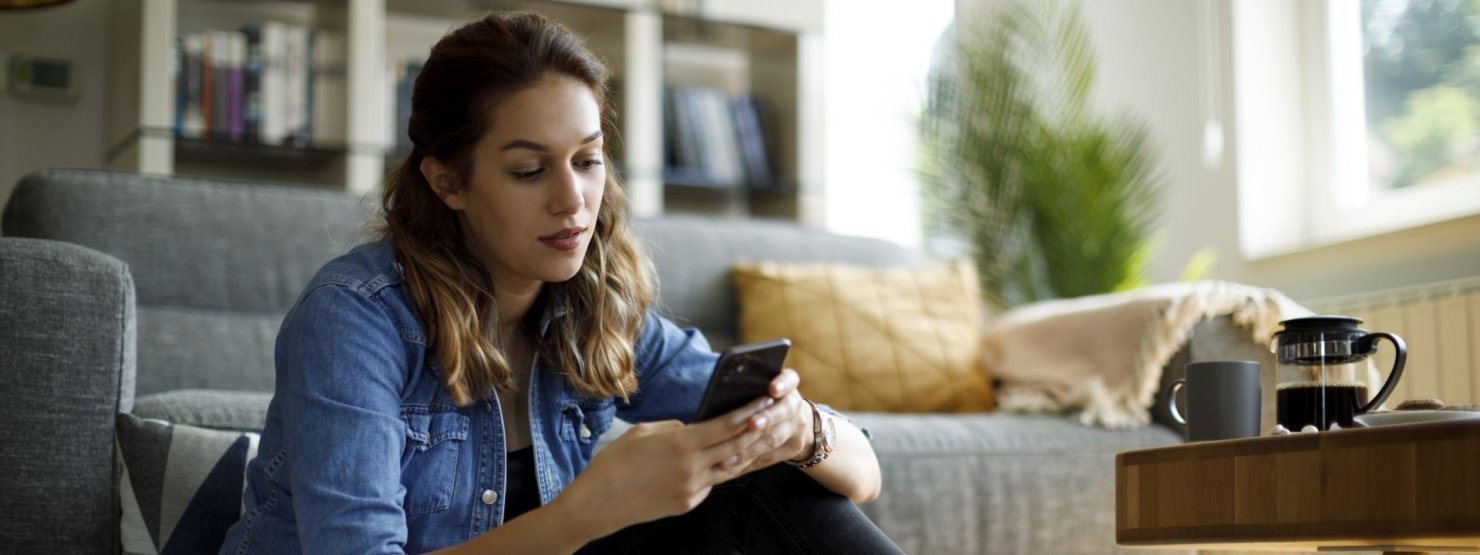 Junge Frau sitzt in ihrem Wohnzimmer auf dem Fußboden und schaut nachdenklich auf ihr Handy.