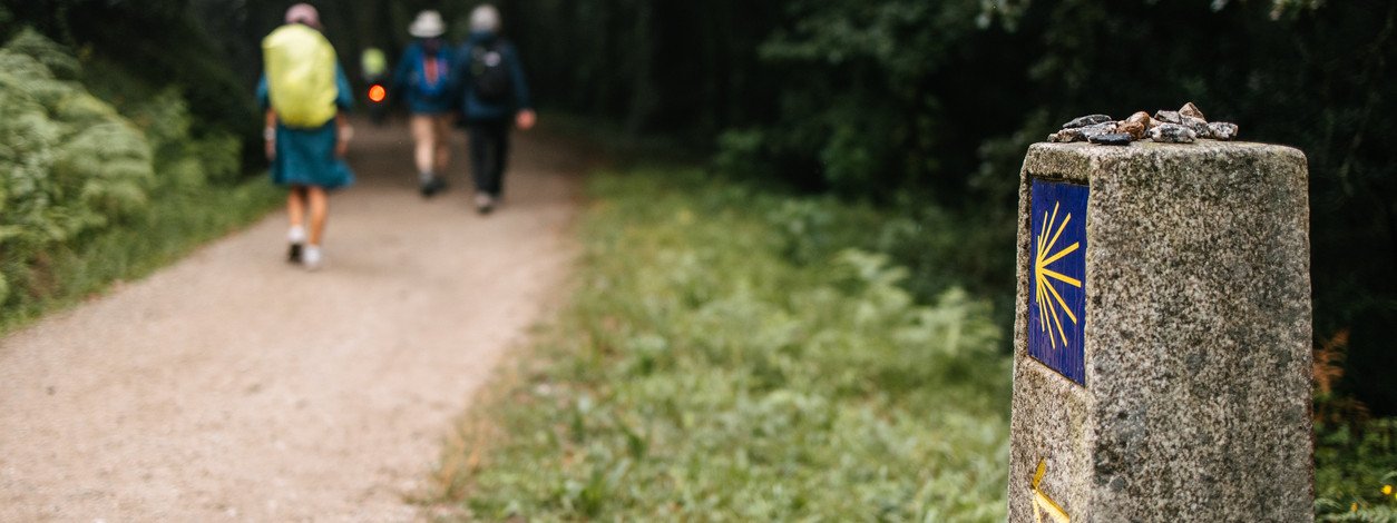Wegmarkierung Jakobsweg, im Hintergrund drei Pilger mit Rucksack und Hut