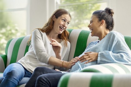 Zwei Frauen, eine davon Schwanger auf einem Sofa. Beide berühren den schwangeren Bauch mit einer Hand.