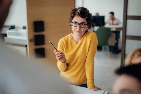 Eine etwa 40 jährige Frau in einem gelben Pullover und mit braunen Locken steht an einem Schreibtisch im Büro. Sie erklährt ihren Kollegen etwas und hebt dabei die Hand. Sie hält einen Stift in dieser Hand.