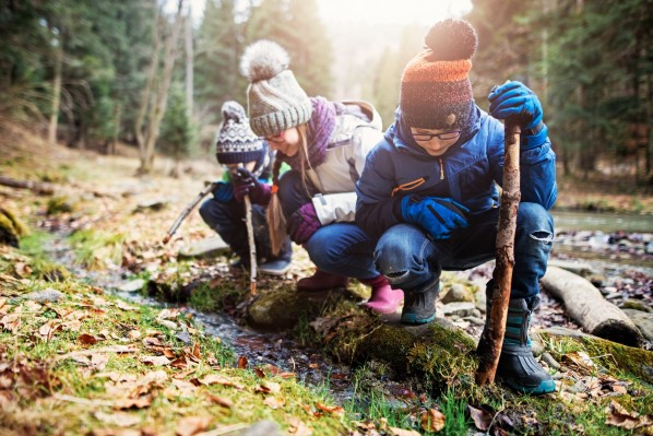 Drei Kinder mit winterlicher Kleidung schauen mit Stöcken in der Hand auf einen kleinen Bach im Wald.