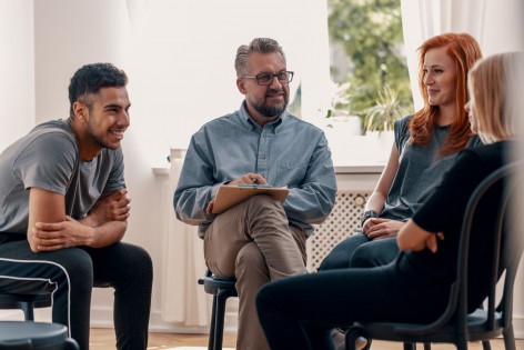 Eine Selbsthilfegruppe sitzt zusammen und unterhält sich. Der Leiter macht sich dabei Notizen. Alle lächeln.