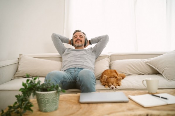 Junger Mann sitzt mit geschlossenen Augen auf der heimischen Couch und hört über Kopfhörer Musik. Sein Hund liegt neben ihm. 