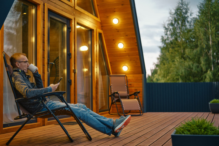 Ein etwa 45-jähriger Mann sitzt auf seiner stark beleuchteten Terrasse, schaut auf sein Smartphone und trinkt aus einer Tasse.