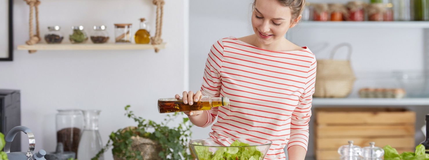 Eine circa 20-jährige Frau steht an ihrer Kücheninsel und gießt etwas pflanzliches Öl auf ihren Salat.