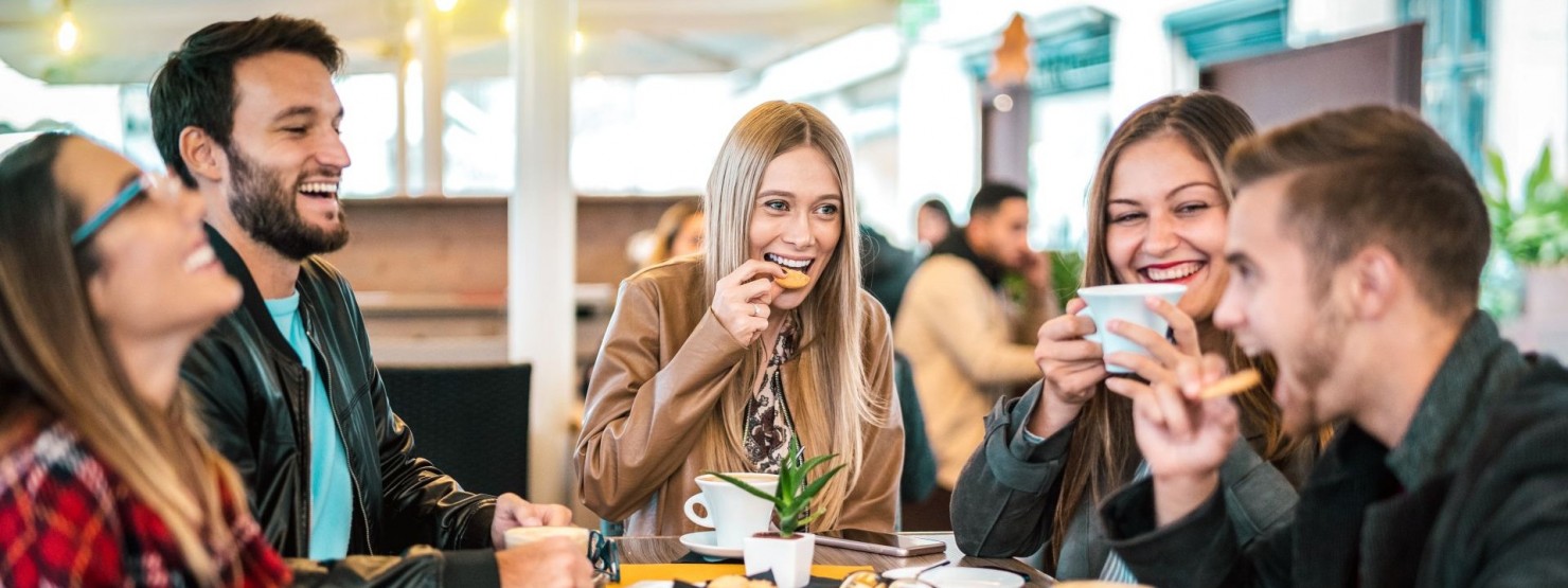 Eine Gruppe von Freunden sitzt im Außenbereich eines Restaurants und lacht ausgiebig. Dabei essen und trinken sie.