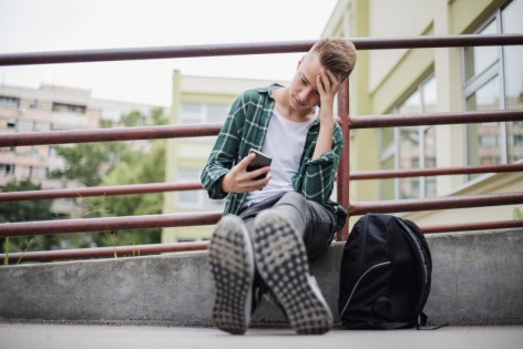 Ein ca. 14-jähriger Junge sitzt vor der schule auf einer Stufe und scha ut traurig auf sein Smartphone