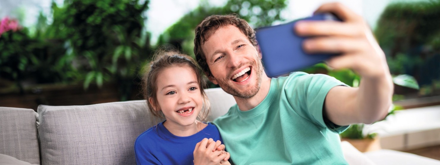 Vater und Tochter sitzen auf der Couch und lächeln breit in die Handykamera.