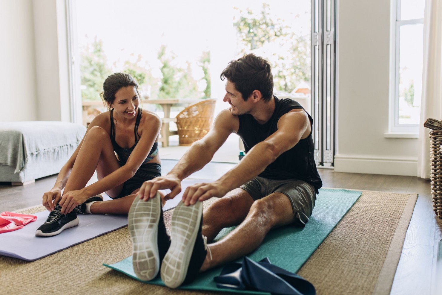 Eine etwa 25 jähriger Frau un ein etwa 25 jähriger Mann sitzen in einem Wohnzimmer auf Yogamatten. Die Frau bindet ihren Sportschuh und der Mann dehnt seine Beine. Beide lächeln sich an und sind verschwitzt.