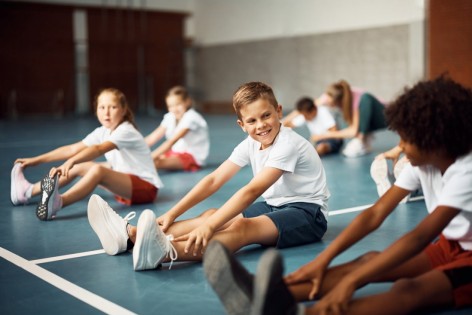Happy elementary student and his classmates stretching on the floor while warming up during exercise class at school gym.