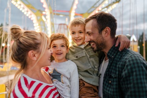 Eine vierköpfige Familie steht lachend an einem Karussell auf dem Jahrmarkt.