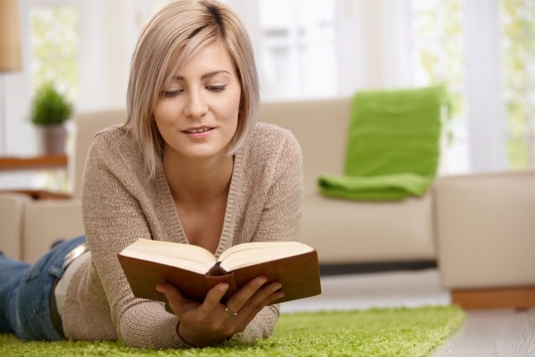 Eine etwa 30 jährige Frau liegt auf dem Boden im Wohnzimmer und liest in einem Buch.