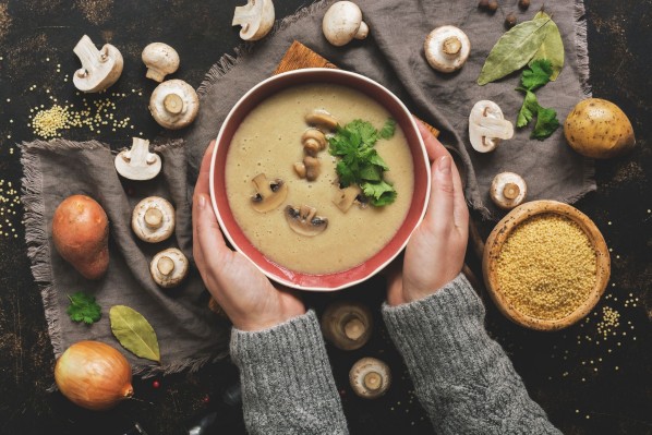 Zwei Hände halten eine Schale mit einer Suppe. In dieser befinden sich Pilze und Koriander. Ringsherum sind auf dem Tisch Pilze, Zwiebeln und Gewürze verteilt.