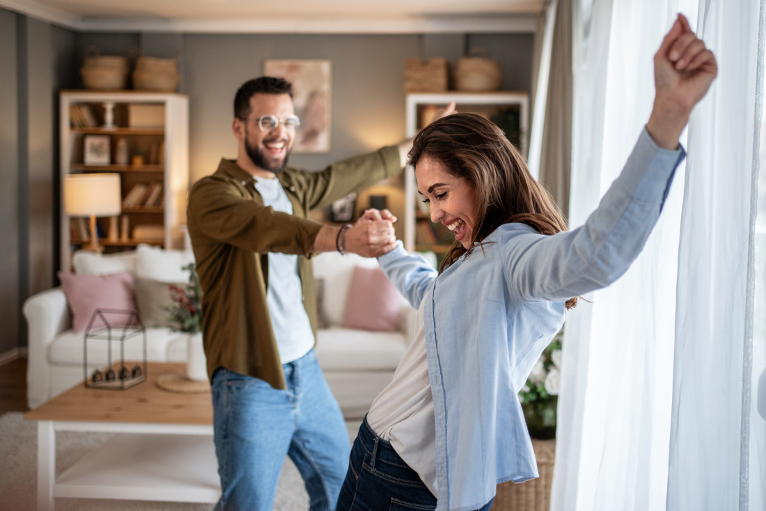 Paar, etwa Mitte 30, tanzt fröhlich miteinander im Wohnzimmer. - AOK Sachsen-Anhalt begleitet mit umfassenden Gesundheitsleistungen für alle Lebenslagen. 