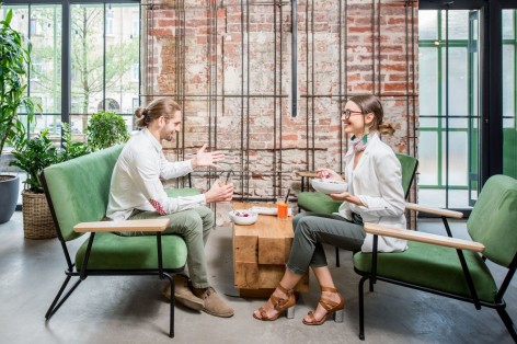 Zwei etwa 25 jährige Personen sitzen jeweils auf zwei Sofas in einem Café. Beide unterhalten sich und essen etwas aus einer Schale.