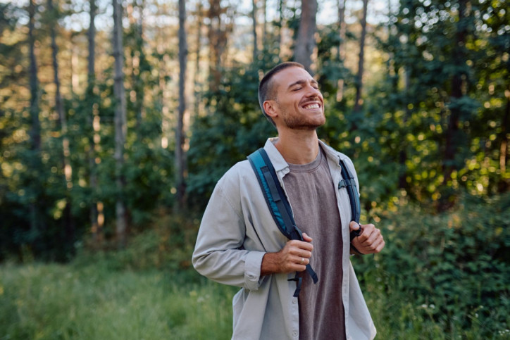 Ein Mann geht im Wald wandern. Er hält kurz inne, hat die Augen geschlossen und lächelt.