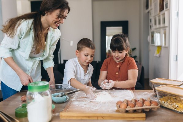 Eine Mutter schaut ihren beiden Kindern beim Kneten eines Teigs zu. Alle lächeln. 