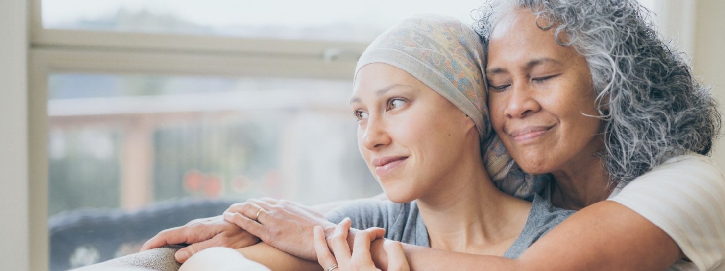 Eine Frau umarmt eine Krebspatientin. Die Frau hat die Augen geschlossen. Die Patientin schaut aus dem Fenster.