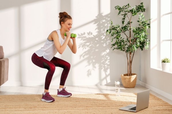 Ein etwa 25 jährige Frau macht in einem Wohnzimmer Sport mit Kurzhanteln. Dabei schaut sie auf einen Laptop.
